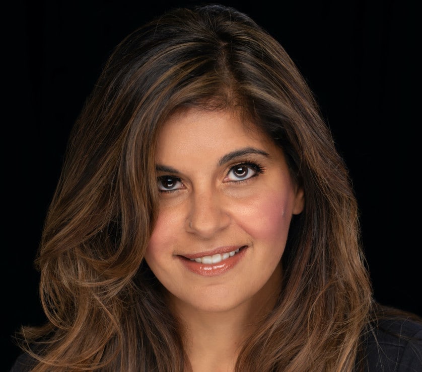 A woman with long, wavy brown hair and brown eyes smiles softly at the camera. She has a natural makeup look and is photographed against a solid black background, reflecting confidence and positivity in women’s sexual health.