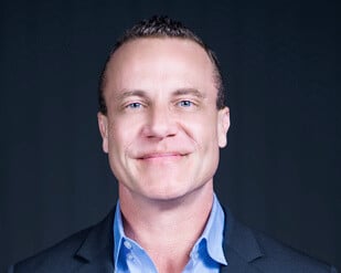 A man with short light brown hair wearing a blue dress shirt and dark blazer, smiling at the camera against a plain dark background, is confident and open about addressing topics like erectile dysfunction.