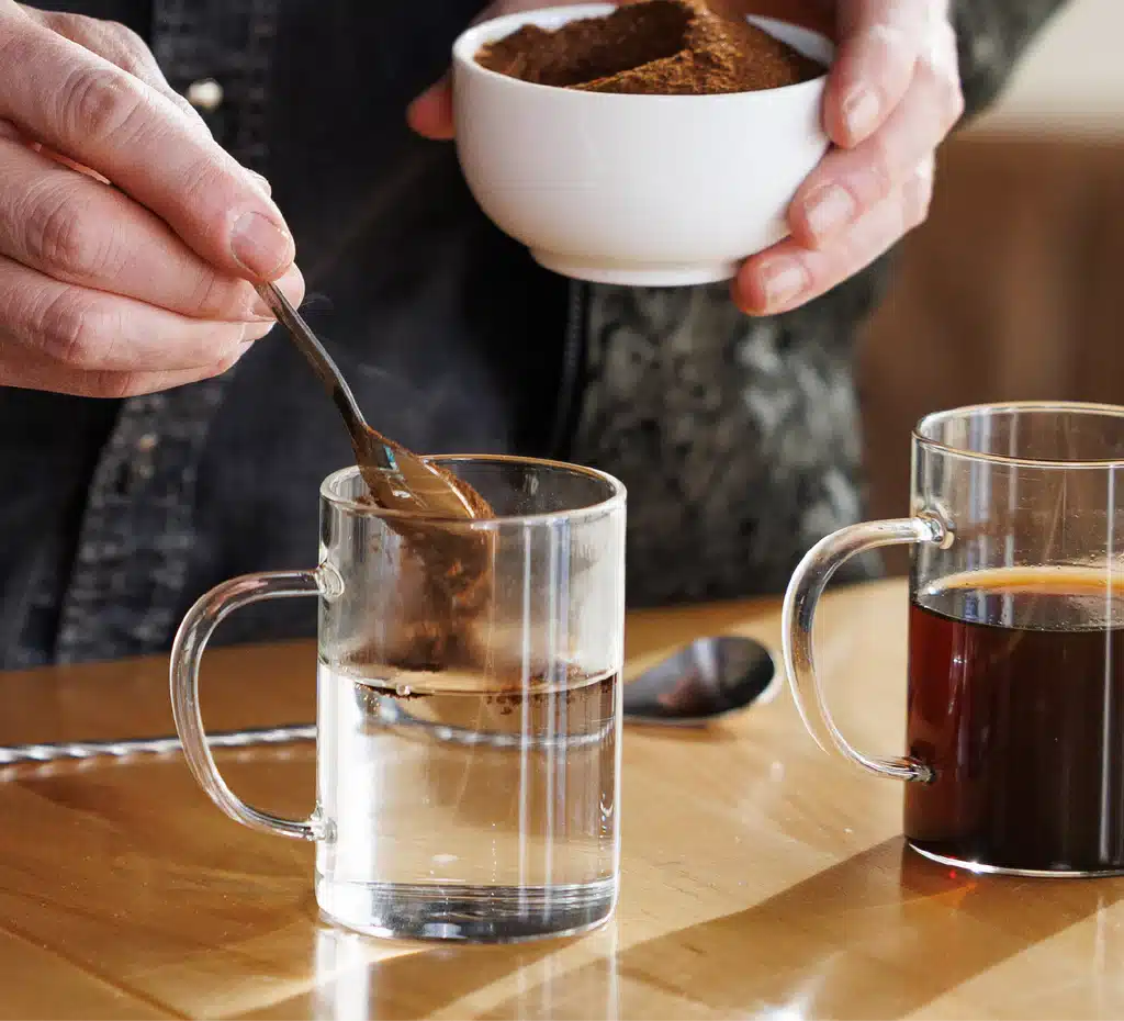 A person adds instant coffee powder from a bowl into a clear glass mug of hot water, with another mug of prepared coffee and a spoon on a wooden table nearby.