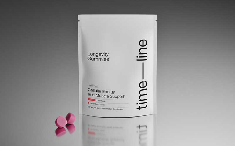 A white pouch labeled Longevity Gummies and time—line stands upright with three pink, round gummies placed in front on a gray surface. The package highlights cellular energy and muscle support.
