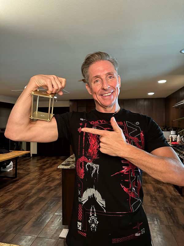 Smiling man in a graphic t-shirt flexes his arm and points to his bicep while holding a small lantern in a kitchen with dark wooden cabinets and flooring.