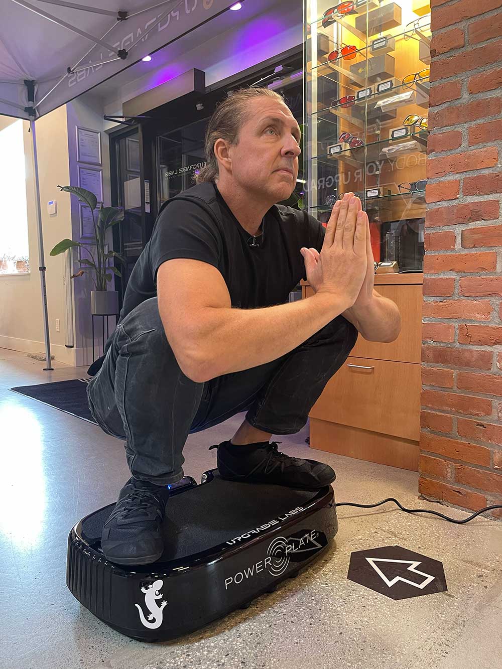 A man squats barefoot on a vibrating Power Plate machine indoors, pressing his palms together in a prayer pose, with shelves and a brick wall in the background.
