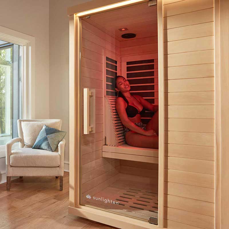 A woman in a bikini relaxes inside a wooden infrared sauna cabin with red lighting. The sauna is in a cozy room with a window, armchair, and throw pillows.