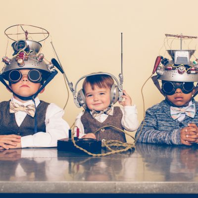 A young boy imagines reading minds of his two friends with a homemade science project. They are dressed in casual clothing, glasses and bow ties. They are serious and sitting at a table with helmets on their heads in front of a beige background. Retro styling.
